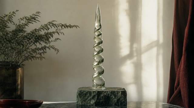 Abstract still life: a tall slender glass spiral tower rising from a stone base, light refracting upward through it like a column